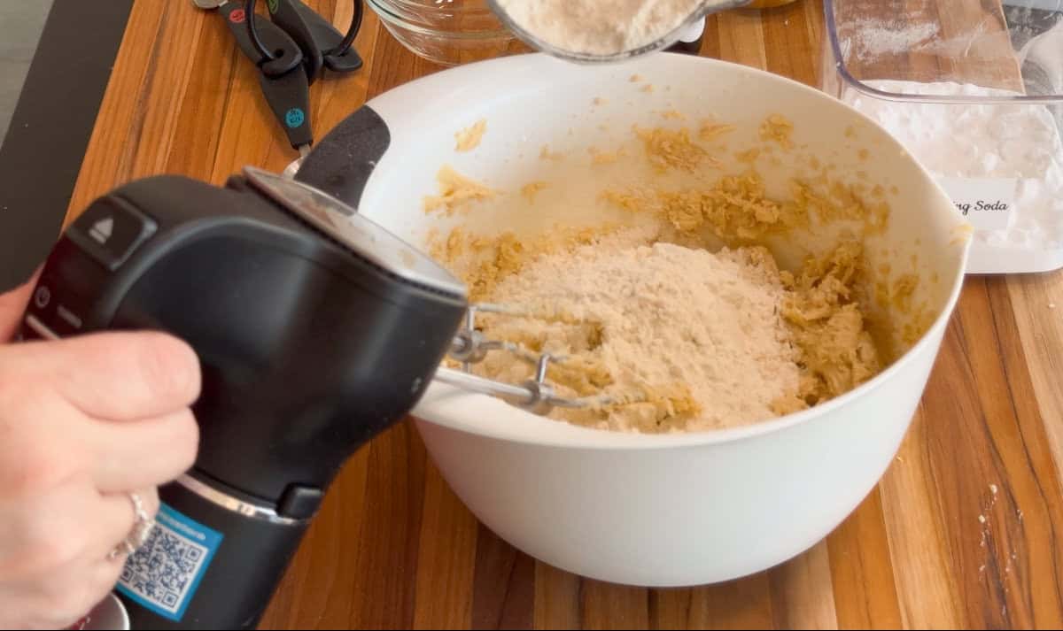 A person uses a hand mixer to blend flour into cookie dough in a large white mixing bowl on a wooden countertop. Measuring spoons, a glass bowl, and a baking soda container are nearby.