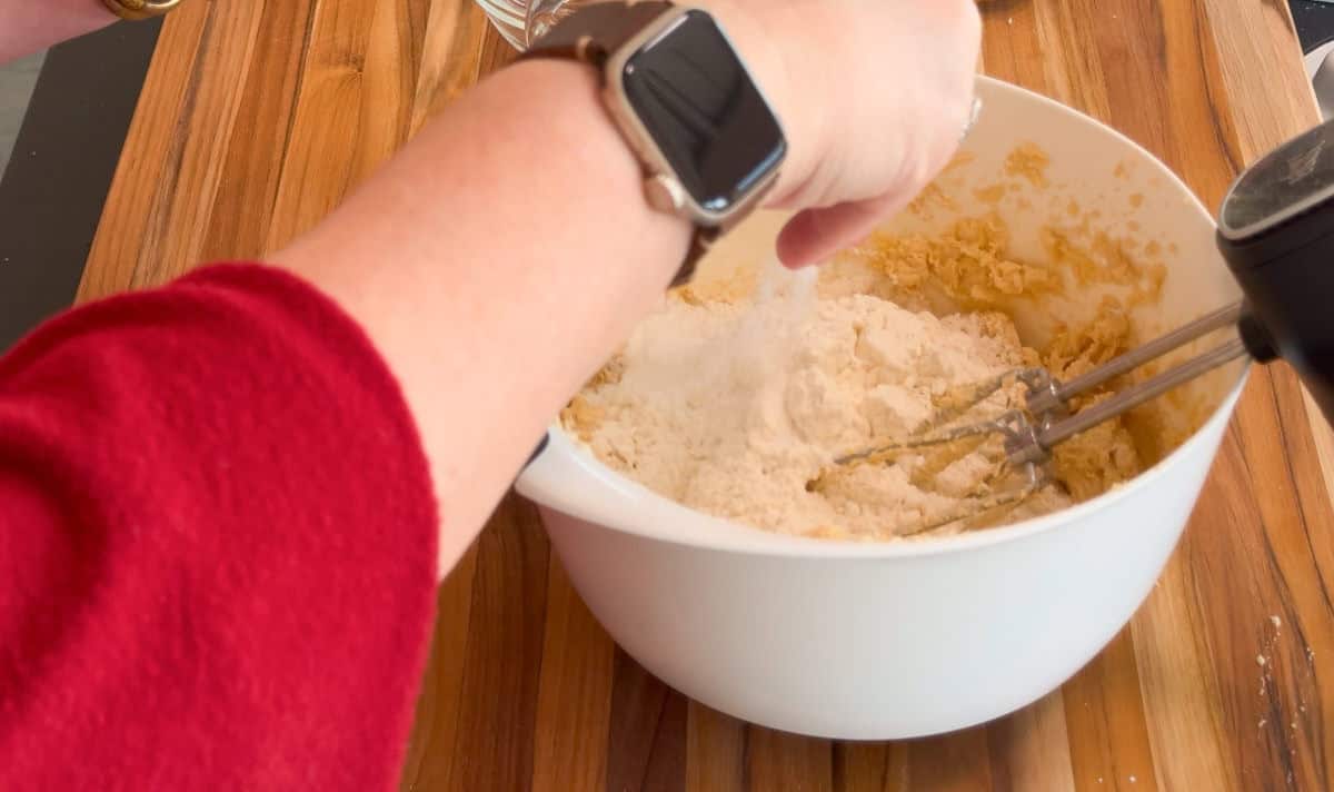 A person wearing a red sleeve and a smartwatch is mixing flour into a dough in a white bowl using an electric hand mixer on a wooden countertop.