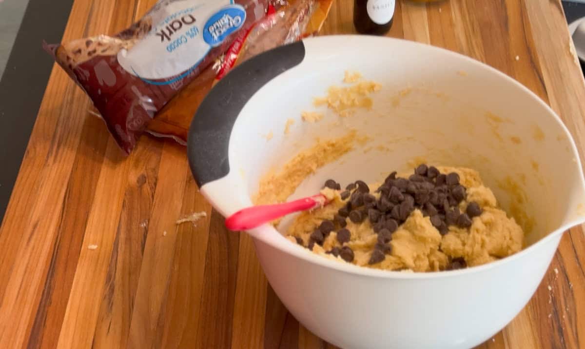 A white mixing bowl with cookie dough and chocolate chips sits on a wooden countertop next to an open bag of chocolate chips. A red spatula is in the dough and a small bottle is in the background.
