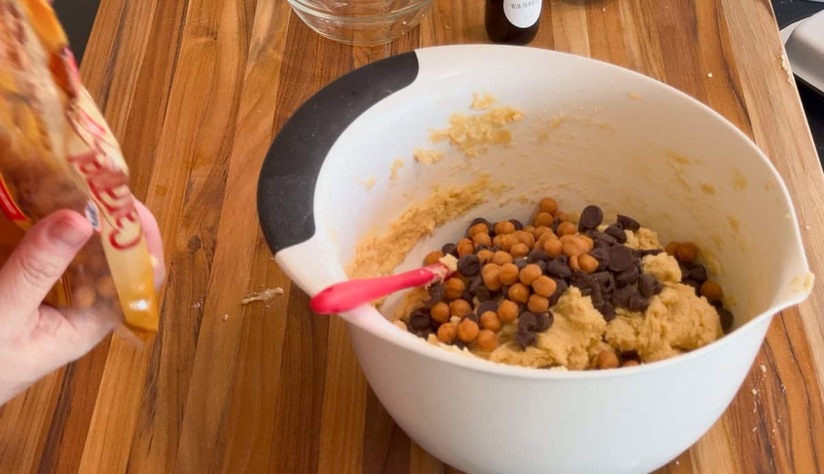 A white mixing bowl with cookie dough, chocolate chips, and caramel chips sits on a wooden countertop. A hand on the left holds an open bag of caramel chips, and a red spatula is in the bowl.