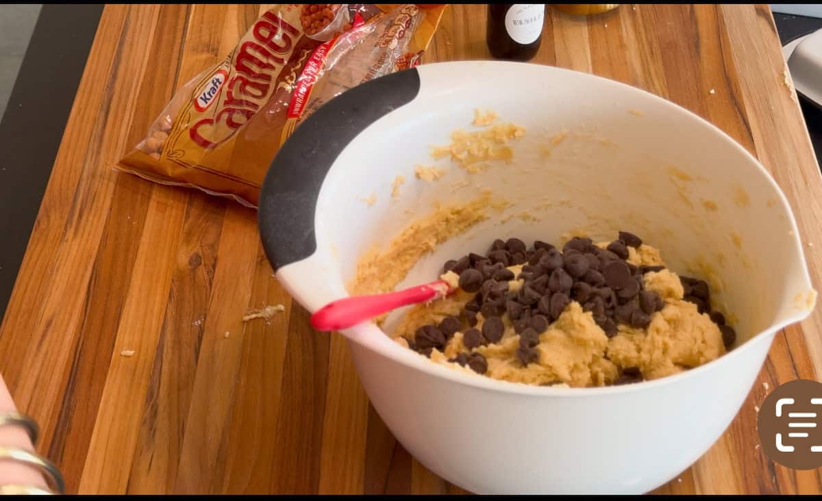 A white mixing bowl with cookie dough and chocolate chips sits on a wooden surface. A red spatula rests in the bowl, and a bag of Kraft caramels is partially visible in the background.