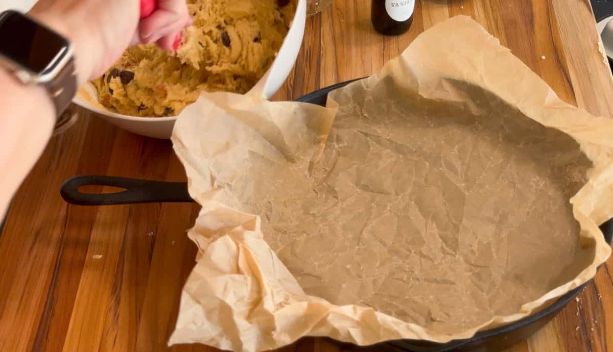 A person holds a bowl of cookie dough above a cast iron skillet lined with crumpled parchment paper on a wooden surface. Some baking ingredients and utensils are partially visible nearby.