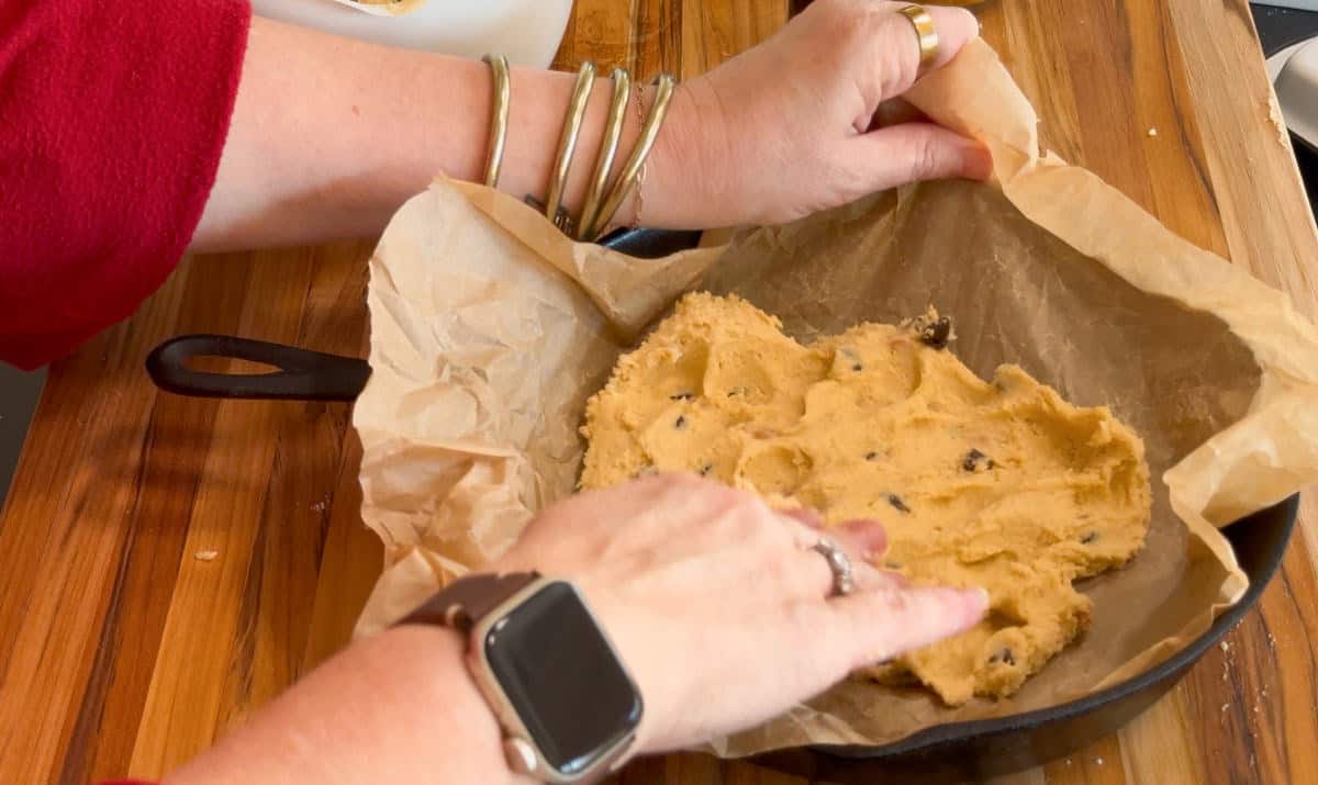 A person presses chocolate chip cookie dough into a cast iron skillet lined with brown parchment paper on a wooden surface. The person's hands, one wearing a smartwatch, are visible spreading the dough.