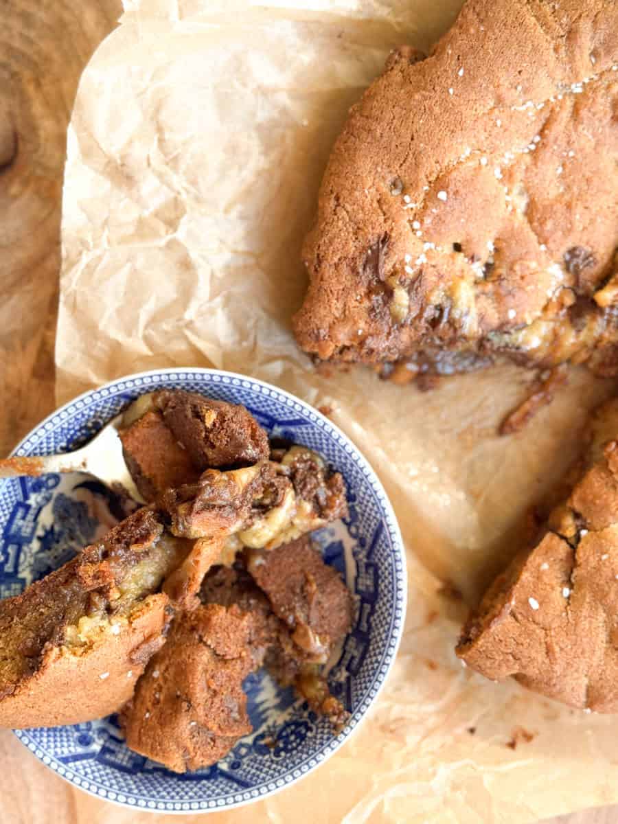 A blue and white patterned bowl filled with pieces of a baked dessert, possibly cookies or blondies with chocolate and nuts, sits on brown parchment paper next to broken larger pieces of the same dessert.