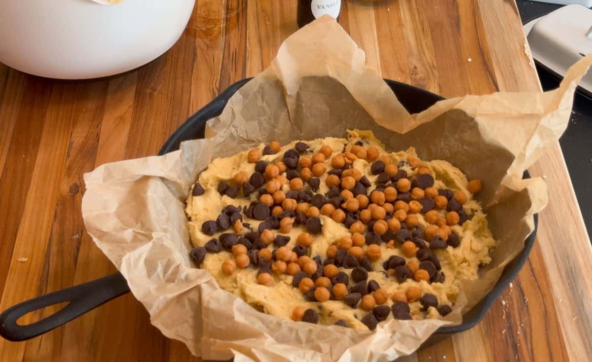 A cast iron skillet lined with parchment paper is filled with cookie dough topped with chocolate chips and butterscotch chips, sitting on a wooden countertop.
