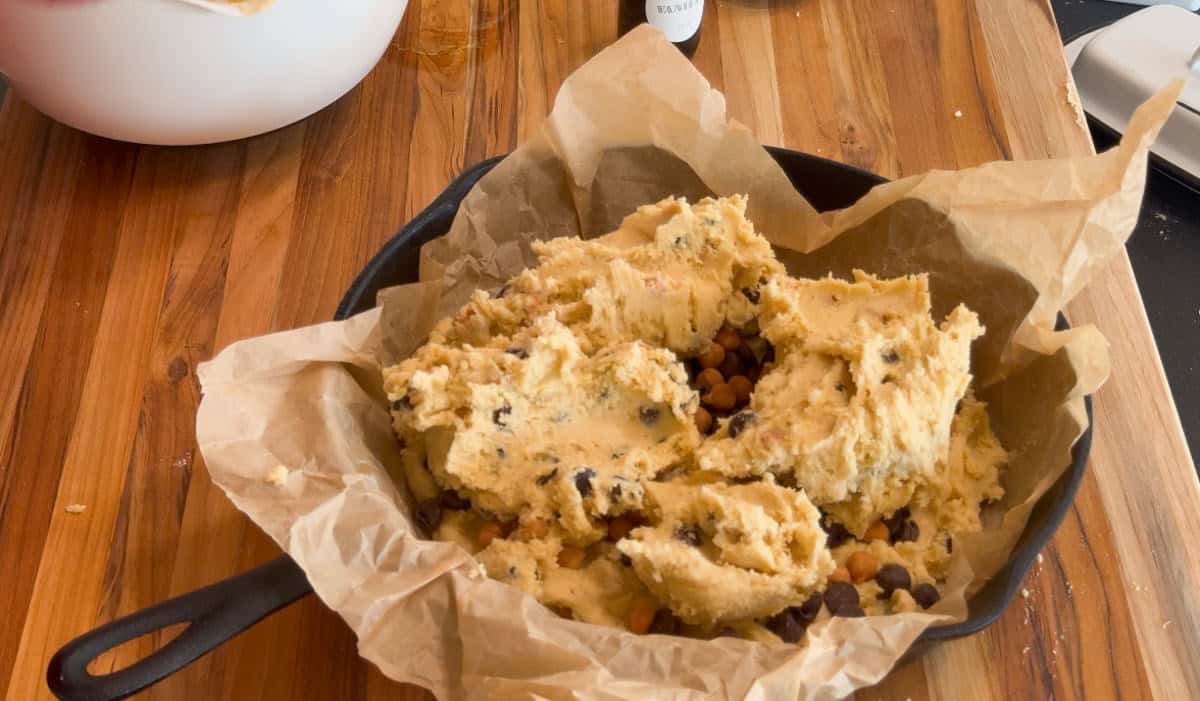 A cast iron skillet lined with brown parchment paper is filled with raw cookie dough mixed with chocolate chips, sitting on a wooden surface.