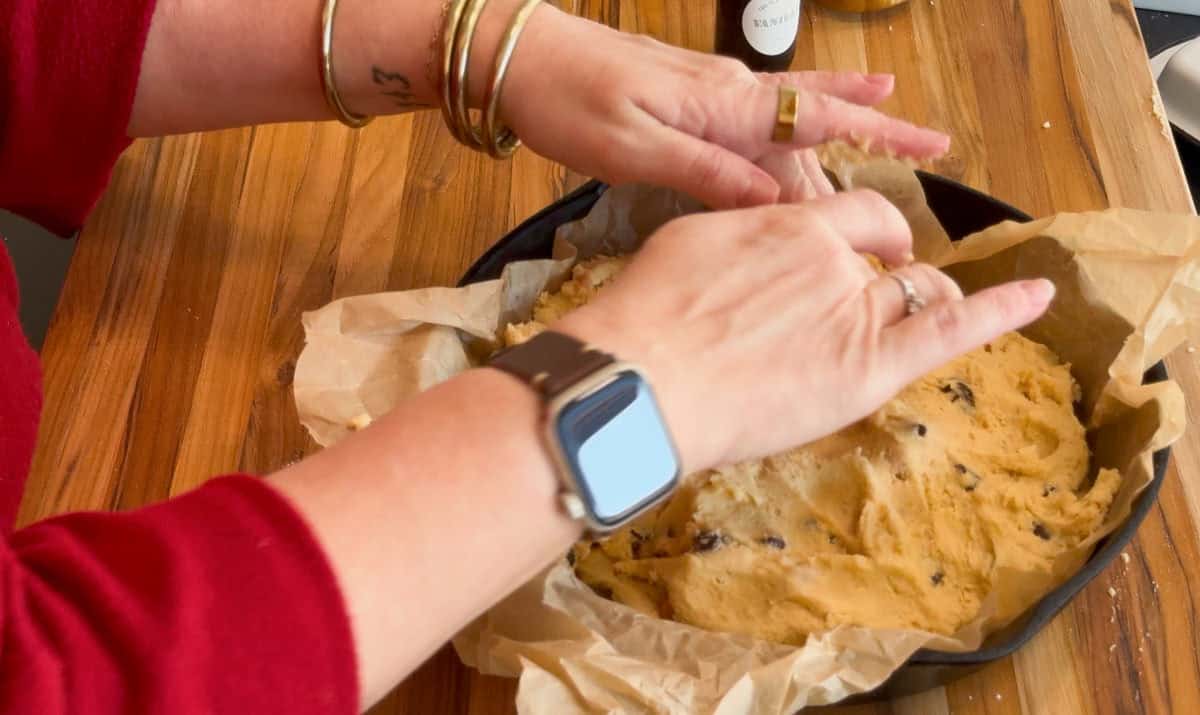 A person wearing a red sleeve and several bracelets presses cookie dough with chocolate chips into a parchment-lined baking pan on a wooden surface.