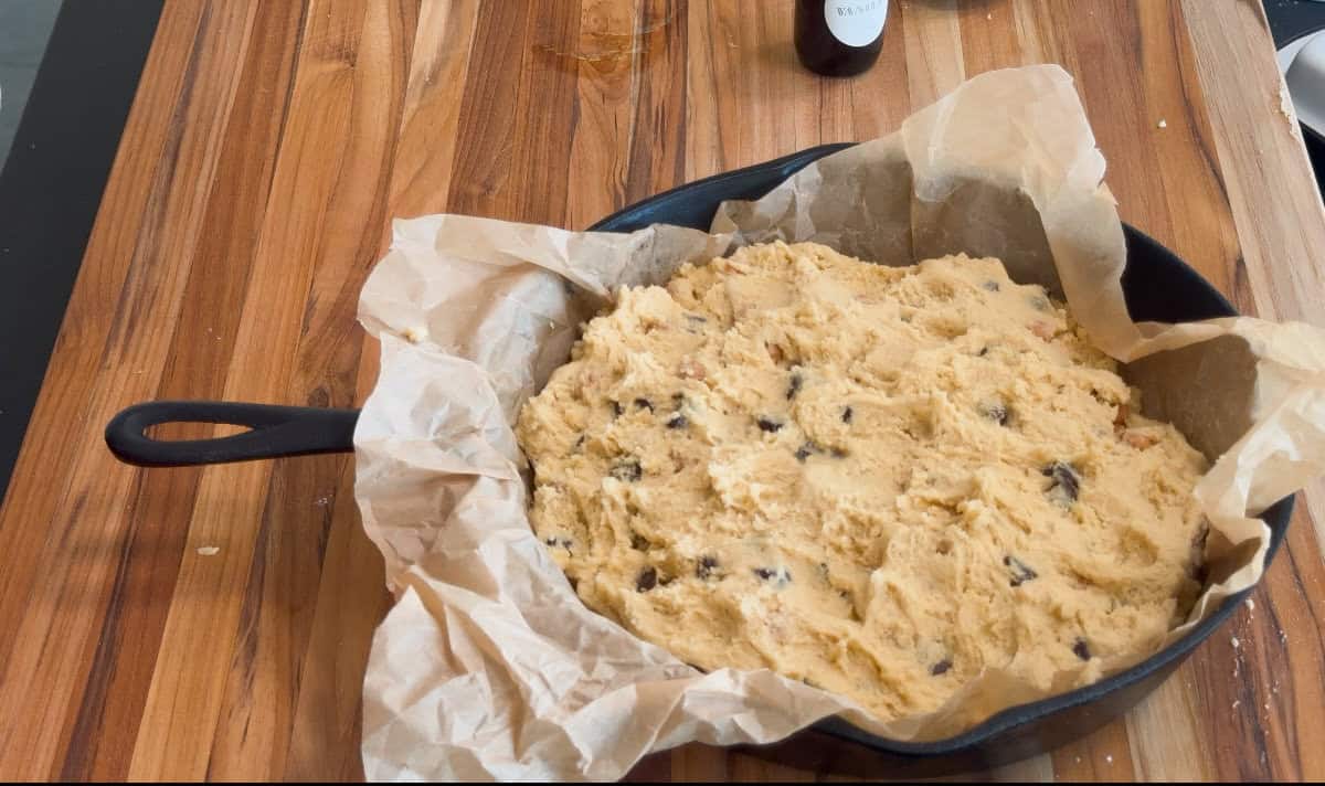 A cast iron skillet lined with parchment paper is filled with unbaked chocolate chip cookie dough, sitting on a wooden countertop. A bottle and some crumbs are visible in the background.