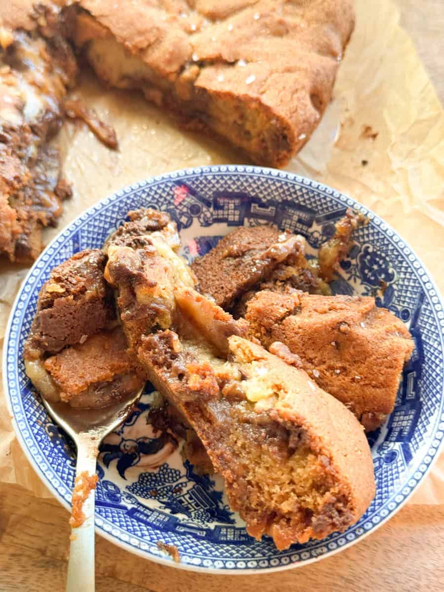 A blue and white patterned bowl holds several slices of a gooey, chocolate and caramel-studded cake. A fork rests in the bowl. In the background, more cake sits on baking paper.