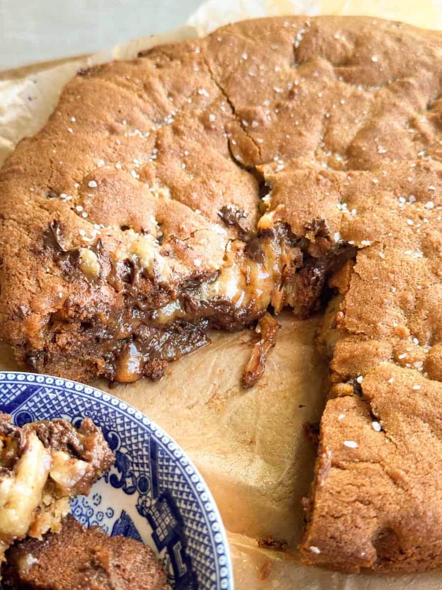 A large, thick cookie pie with a gooey chocolate and caramel filling sits on parchment paper. One slice has been cut out and placed on a blue and white patterned plate in the foreground.