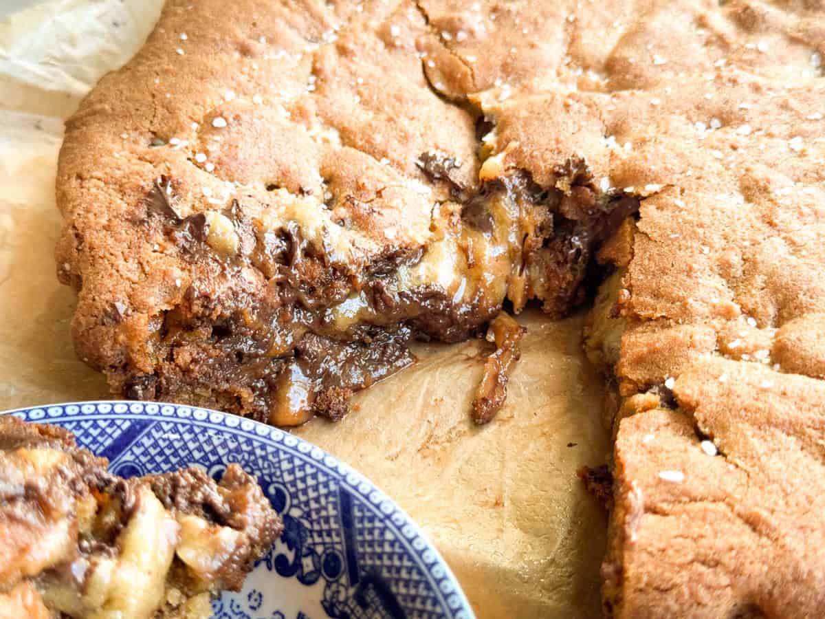 Close-up of a large, gooey chocolate chip cookie on parchment paper with a wedge removed; a piece of the cookie sits on a blue and white patterned plate in the foreground.