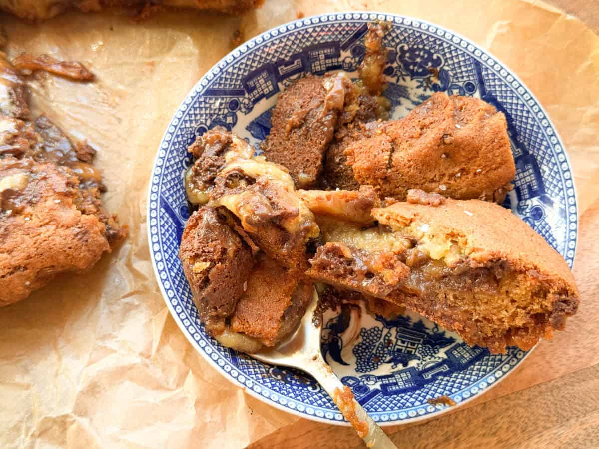 A blue and white patterned bowl holds chunks of gooey, golden-brown dessert with melted chocolate and caramel. A spoon rests in the bowl. The bowl sits on parchment paper with more dessert visible nearby.