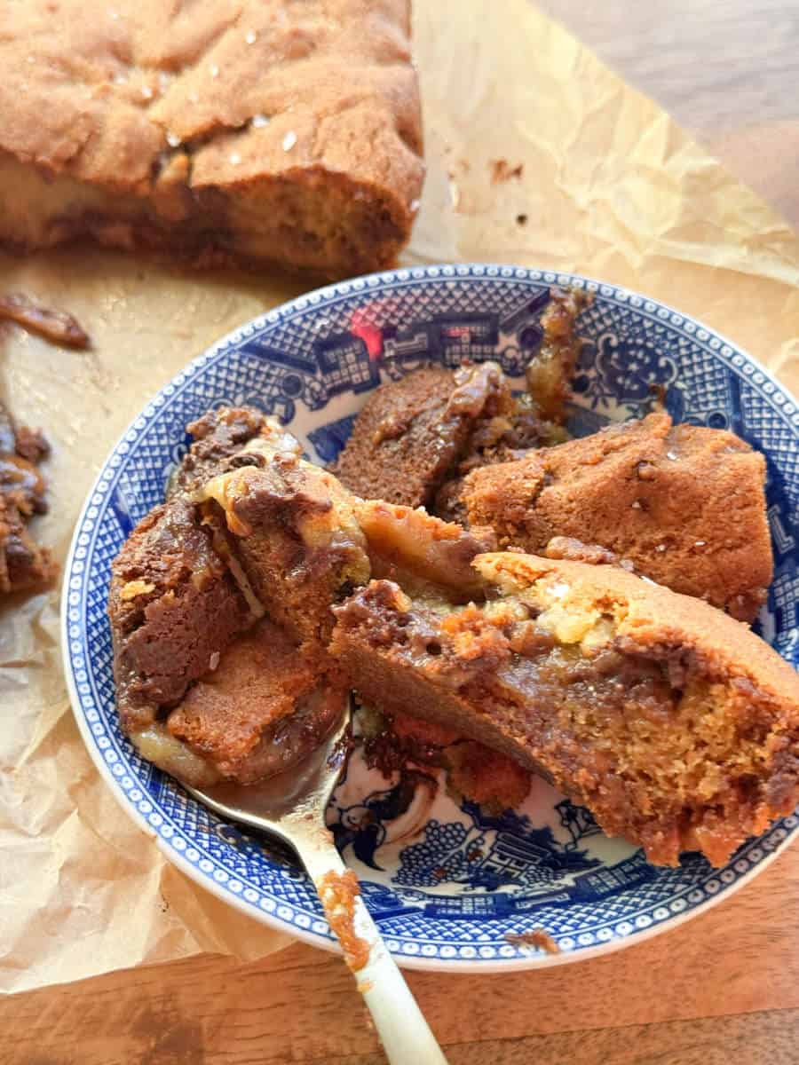A blue and white patterned bowl contains pieces of gooey chocolate chip cookie cake on a wooden surface. A spoon rests in the bowl. More cookie cake sits on parchment paper in the background.