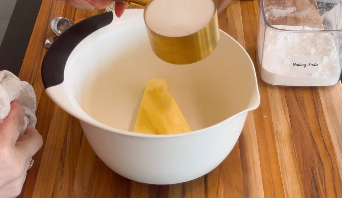 A person holds a measuring cup of sugar above a white mixing bowl containing two sticks of butter on a wooden countertop. A container labeled "Baking Soda" sits nearby.