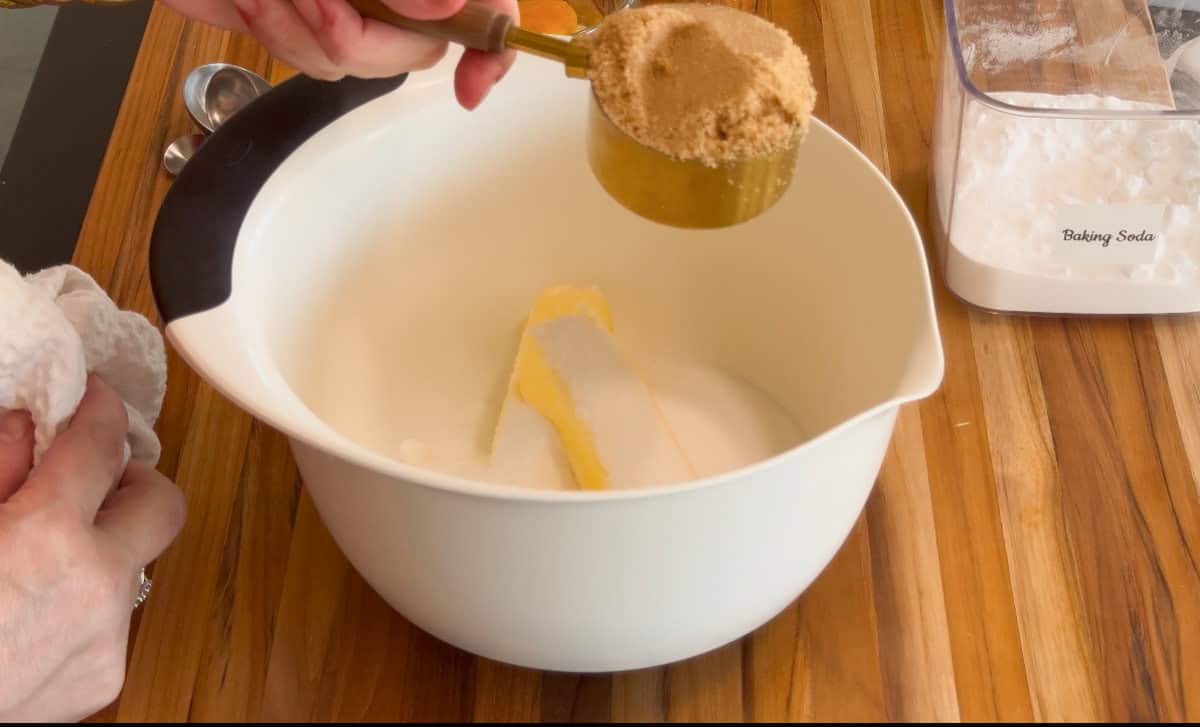 A person adds a measuring cup of brown sugar to a white mixing bowl containing butter and white sugar on a wooden surface. A container labeled "Baking Soda" is nearby.