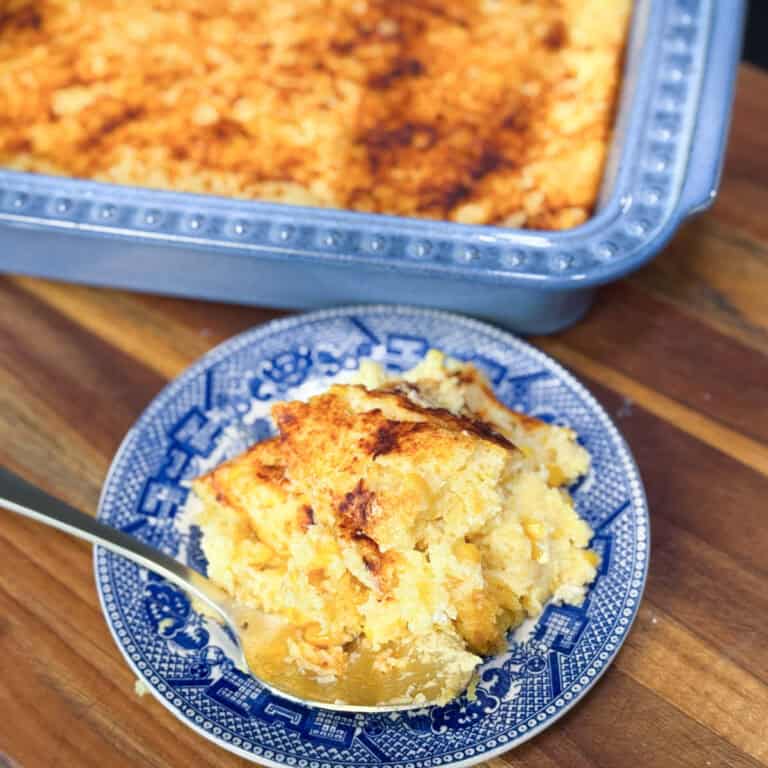 A serving of baked corn casserole on a blue and white plate with a spoon, placed in front of a metal baking dish on a wooden surface.