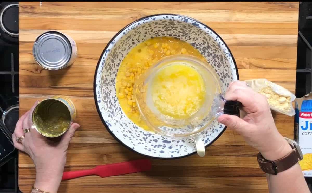 A person pours melted butter into a bowl containing eggs and corn while holding an opened can of corn. A red spatula, can lid, and cornbread mix packet are on the wooden countertop.