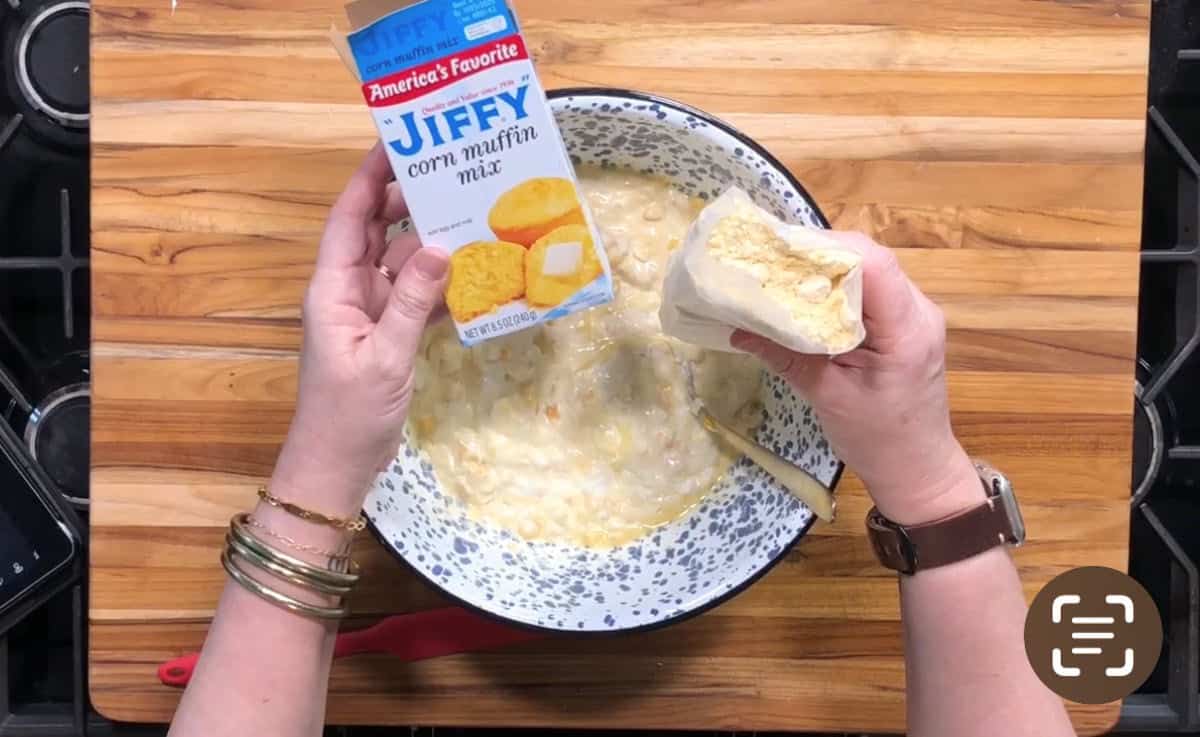 A person is holding a box and pouch of Jiffy corn muffin mix over a bowl of partially mixed batter on a wooden surface. The person is wearing bracelets and a watch.