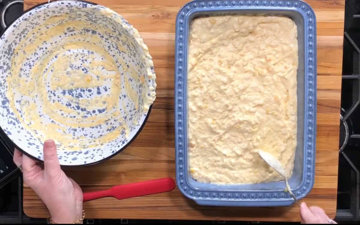 A person holds an empty mixing bowl with batter residue in one hand and uses a spoon to spread batter evenly in a rectangular baking dish on a wooden surface. A red spatula rests nearby.