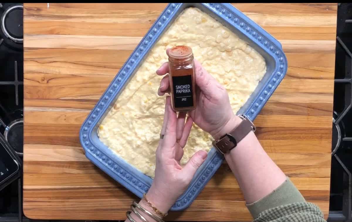 A person holds a small jar labeled “Smoked Paprika” above a blue baking dish filled with an uncooked, creamy mixture on a wooden countertop.