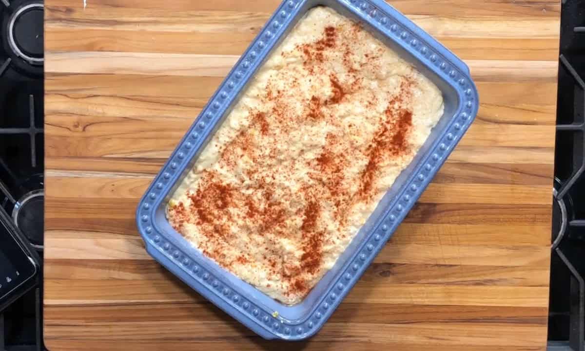 A blue rectangular baking dish filled with a creamy casserole topped with a layer of red paprika, placed on a wooden cutting board.