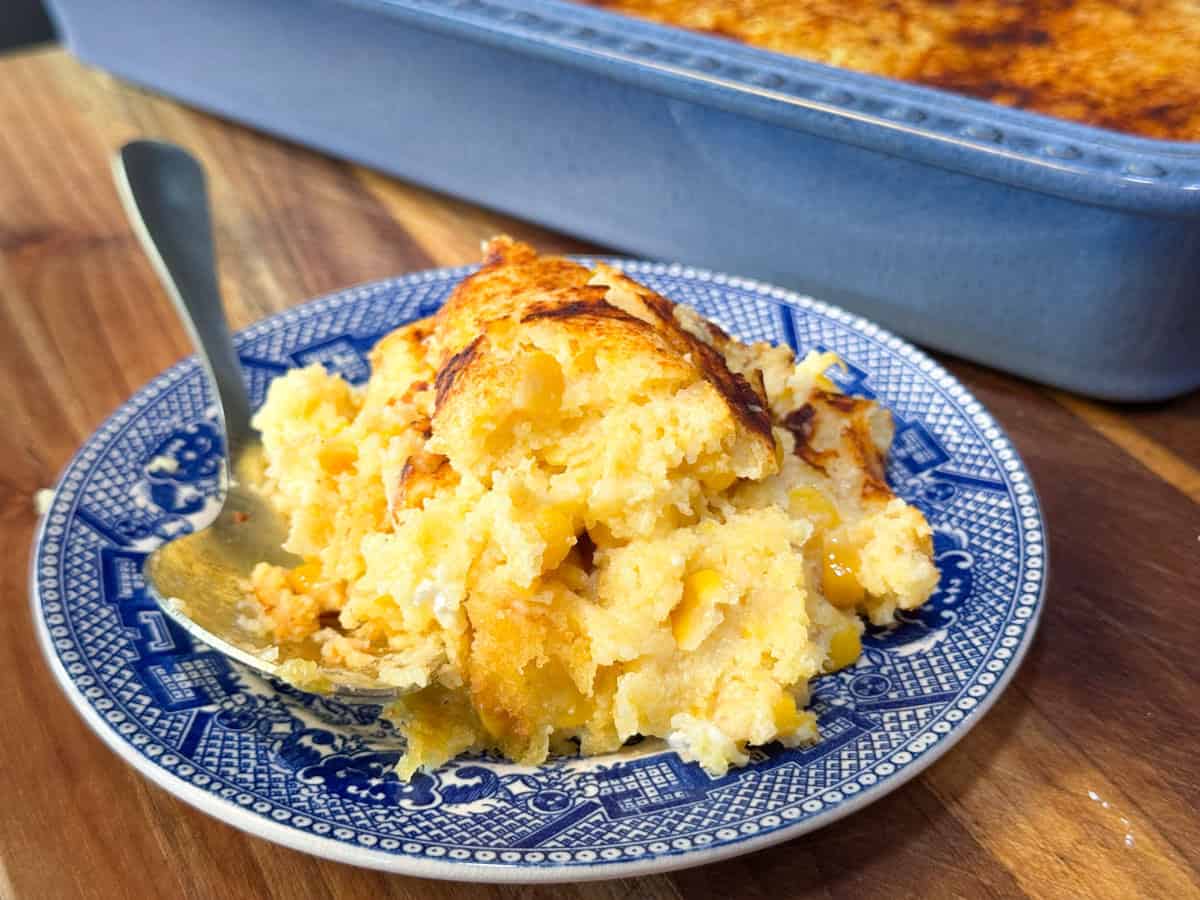 A serving of baked corn pudding on a blue and white patterned plate with a spoon, in front of a blue baking dish on a wooden table.