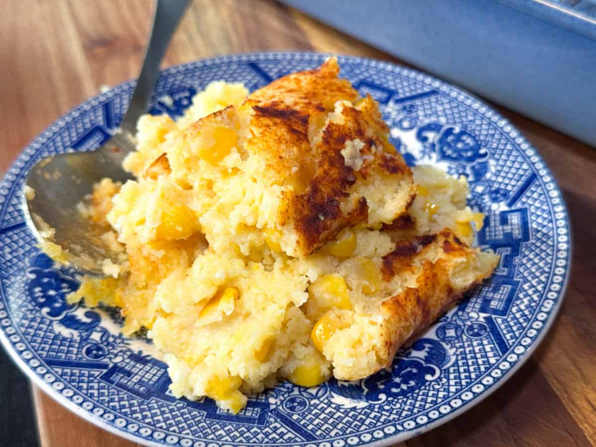 A serving of baked corn casserole with visible corn kernels on a blue and white patterned plate, with a serving spoon beside the food.