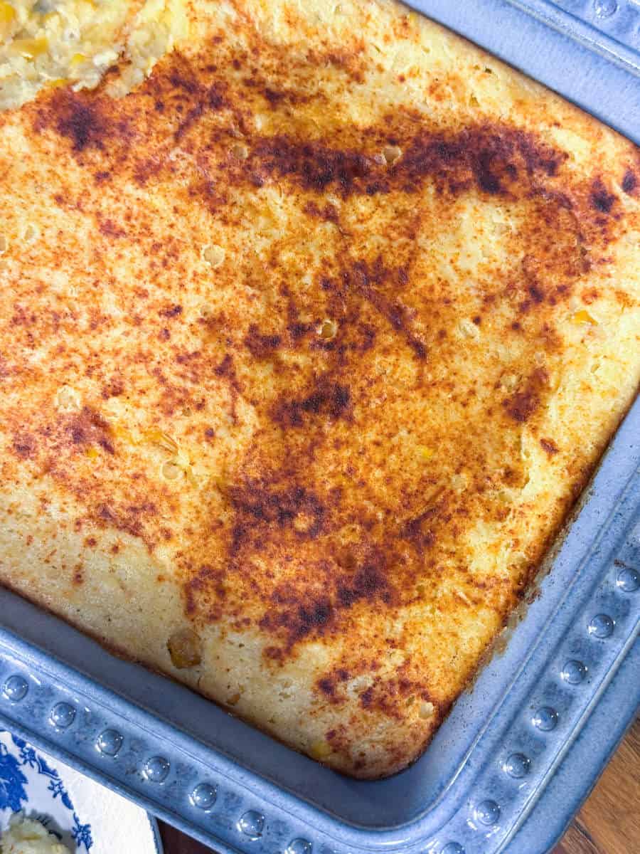 A close-up view of a baked cornbread casserole in a metal baking dish, showing a golden brown, slightly crispy top with some darker browned areas. A portion is missing from the upper left corner.