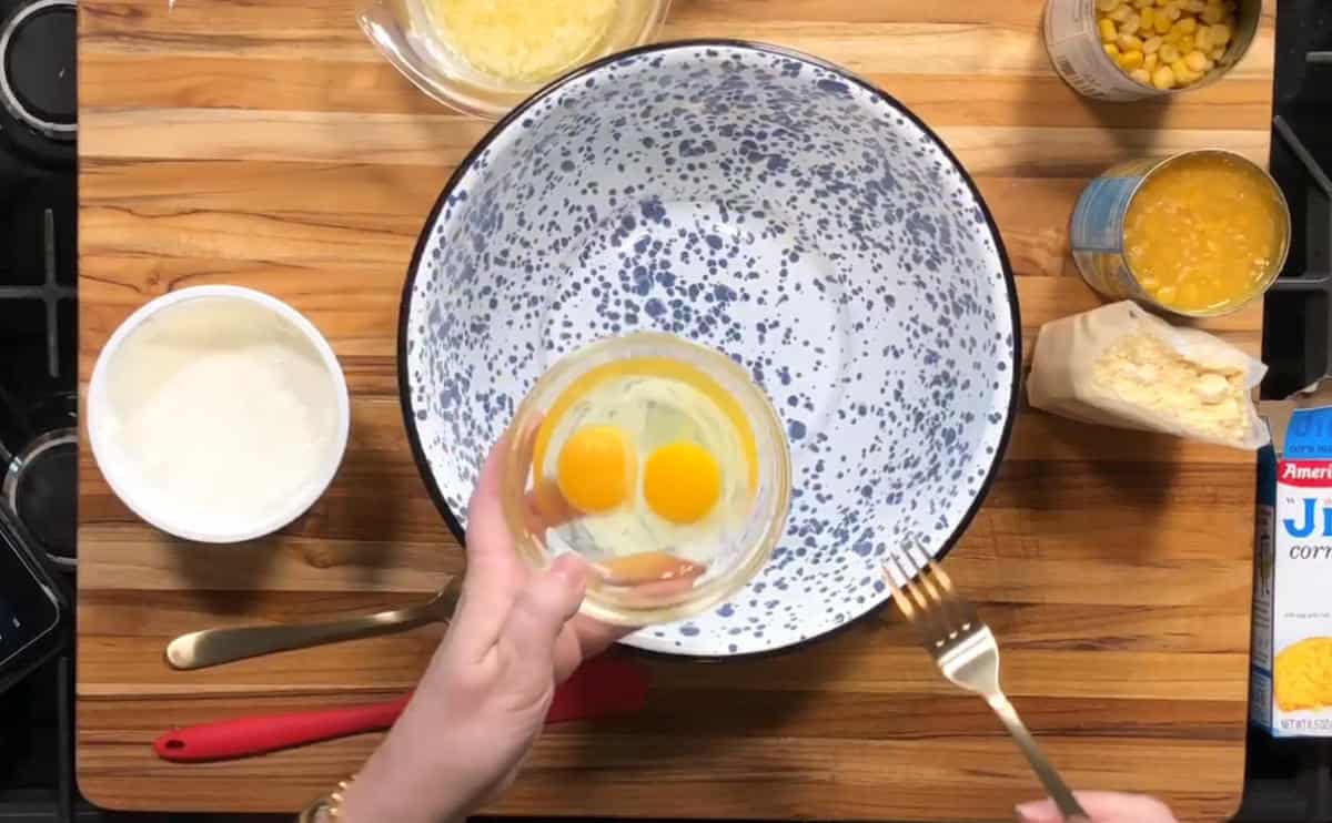 A person holds a small bowl with two cracked eggs above a large speckled mixing bowl on a wooden surface, surrounded by ingredients including sour cream, canned corn, shredded cheese, and a box of cornbread mix.