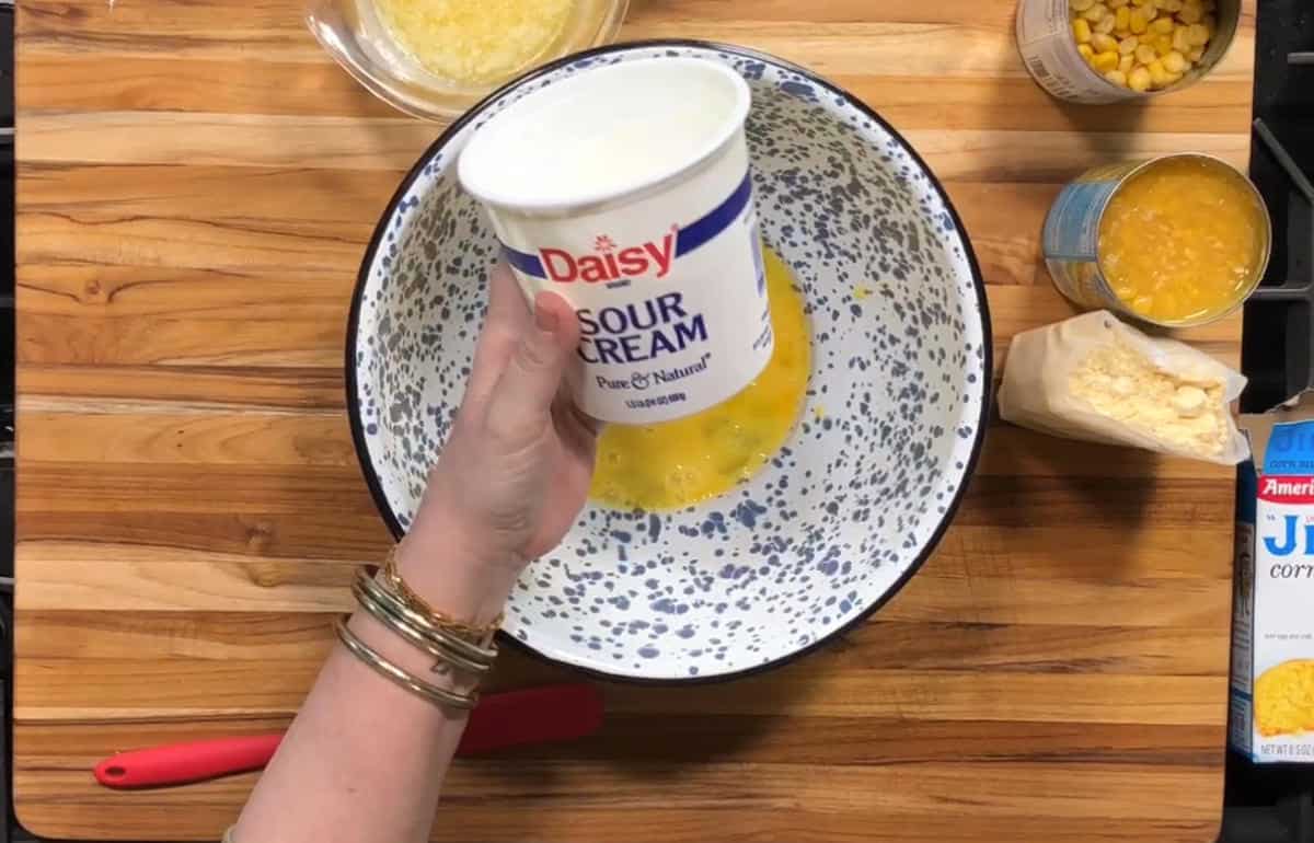 A hand holding a container of Daisy sour cream over a speckled mixing bowl filled with beaten eggs; nearby are opened cans of corn, melted butter, a red-handled spatula, and a box of Jiffy corn muffin mix.