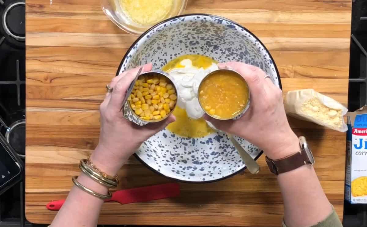 A person holds a can of whole kernel corn and a can of creamed corn over a mixing bowl containing a yellow mixture, with Jiffy corn muffin mix, a spatula, and a bowl of grated cheese nearby.