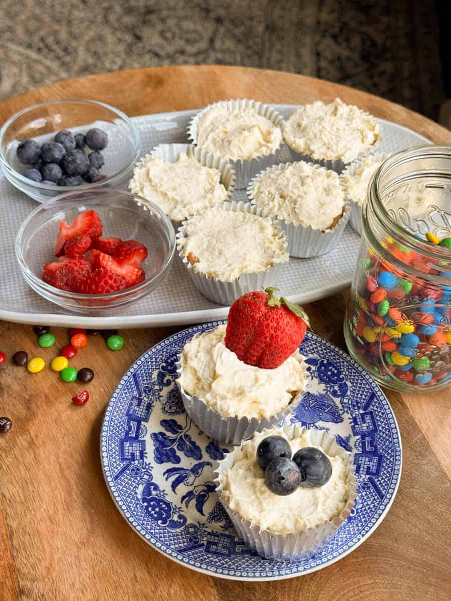 Several cupcakes with white frosting are displayed on plates. Some cupcakes are topped with strawberries or blueberries. A jar of colorful candy and small bowls of fruit are next to the cupcakes on a wooden table.