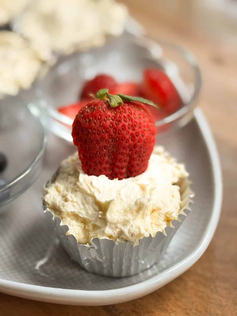 A cupcake in a silver wrapper topped with a large strawberry and white frosting sits on a plate next to a small glass bowl containing whole strawberries.