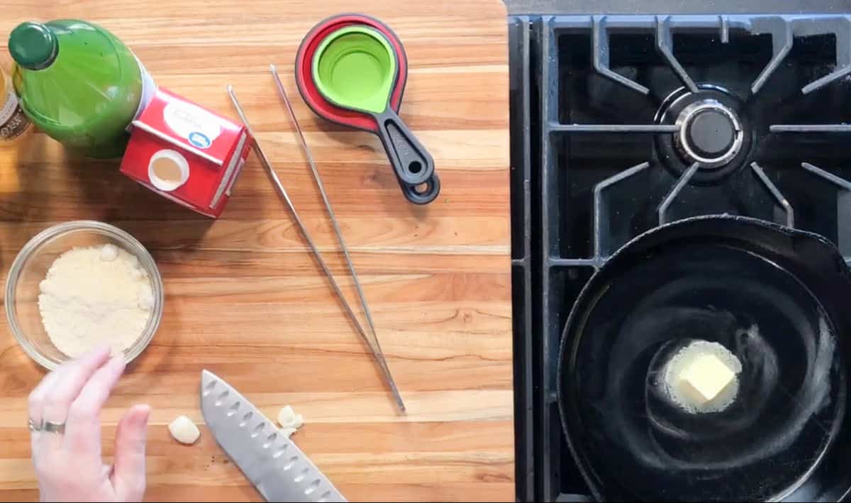 A wooden countertop with flour, garlic, a knife, measuring cups, chopsticks, and a carton of cream beside a stove with a cast iron pan melting butter. A hand is reaching for garlic on the left side of the image.