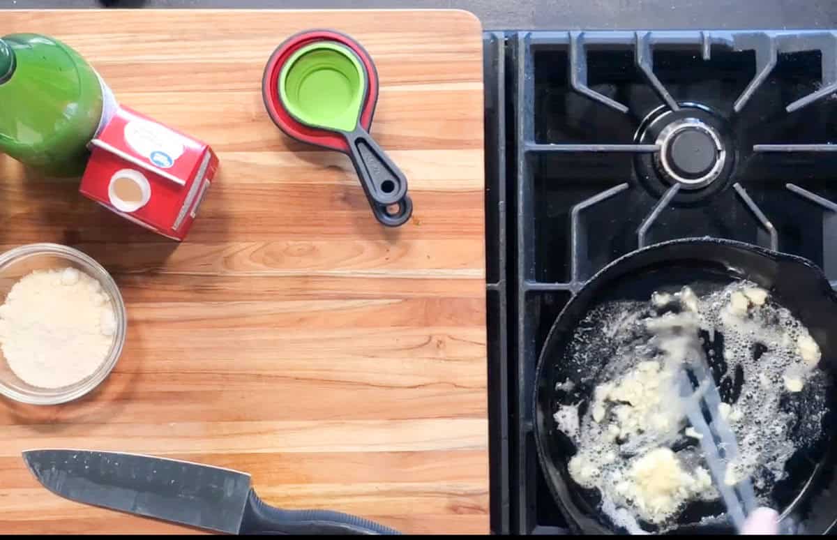 A person stirs a mixture in a black skillet on a gas stove. Nearby on a wooden countertop are a knife, measuring cups, a bowl of flour, a green bottle, and a carton of cream.