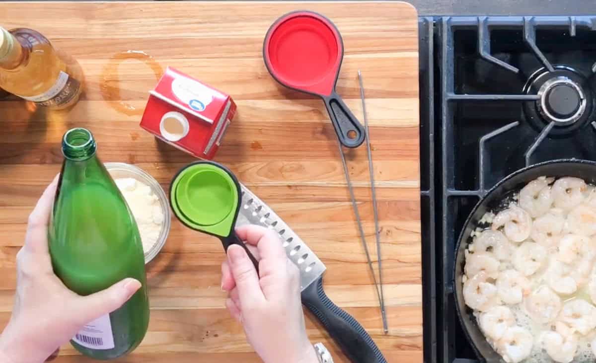 A top view of a kitchen counter with a cutting board holding a bottle, measuring cups, a carton of cream, a bowl of grated cheese, and a knife. On the stove, shrimp are cooking in a pan. Hands hold a green measuring cup and bottle.