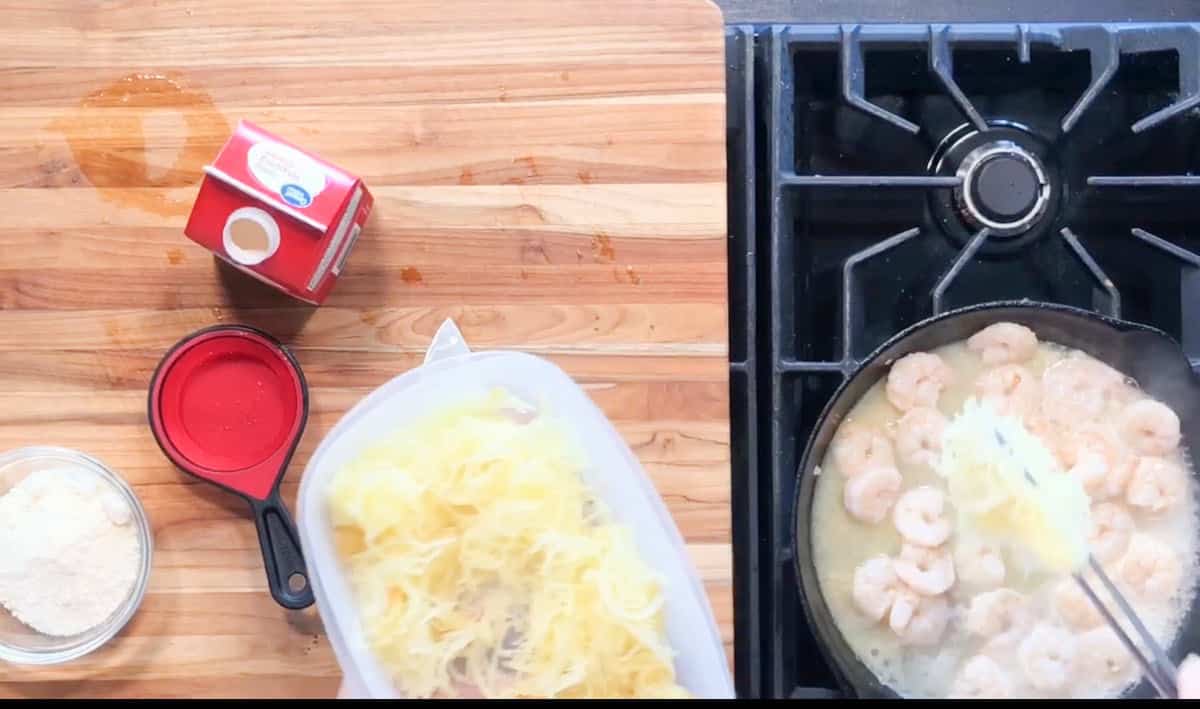 A container of spaghetti squash is held over a skillet of shrimp cooking in a creamy sauce on a stovetop. On the wooden counter are a carton of cream, a small glass bowl of grated cheese, and a red measuring cup.