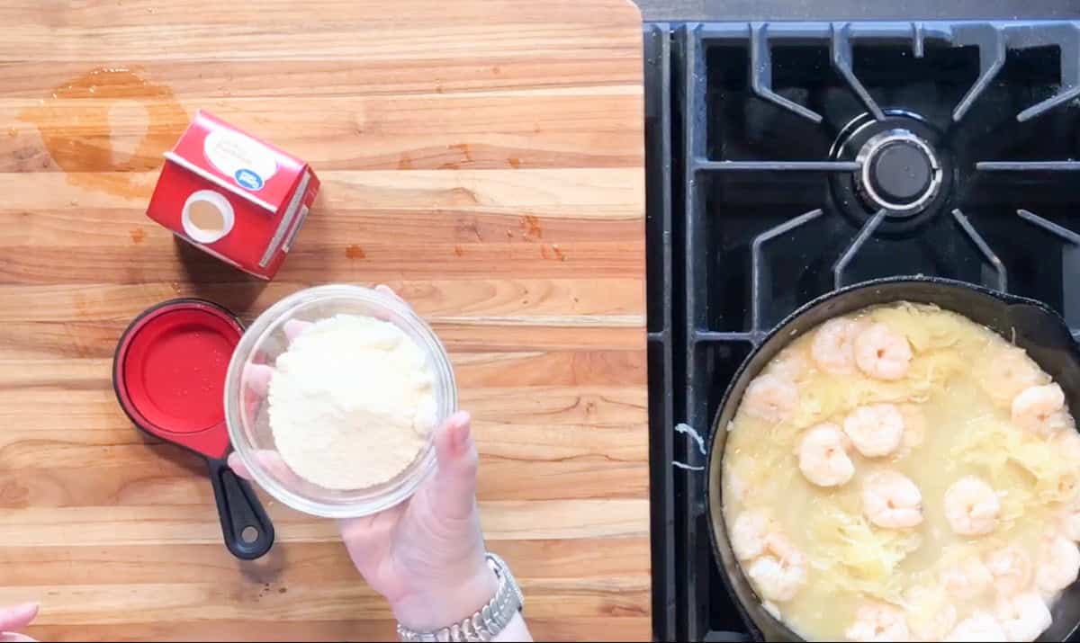A person holds a small glass bowl of grated cheese over a wooden countertop next to a red measuring cup and a carton of cream, with a pan of shrimp and spaghetti cooking on a stove.