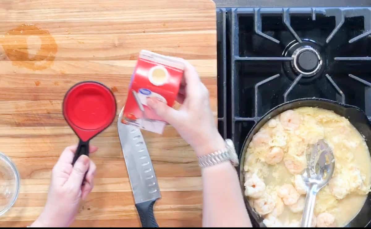 A person holds a red measuring cup and a carton of heavy cream over a wooden cutting board next to a chef’s knife; a pan with shrimp and sauce sits on a stovetop nearby.