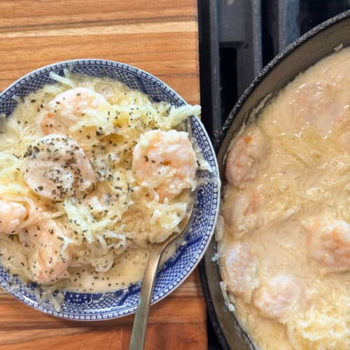 A blue and white plate holds a serving of creamy shrimp and spaghetti squash, topped with herbs. Beside it, a skillet contains more of the same shrimp and squash mixture in sauce. A spoon rests on the plate.