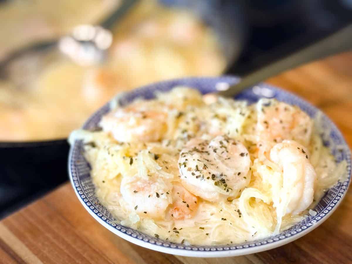 A bowl of spaghetti topped with shrimp and sprinkled with herbs sits on a wooden surface, with a serving spoon and pan of more pasta blurred in the background.