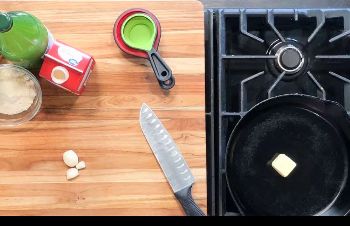A wooden cutting board with garlic cloves, a chef's knife, grated cheese, tomato puree, measuring spoons with oil, and a stove with a cast iron pan containing a pat of butter.