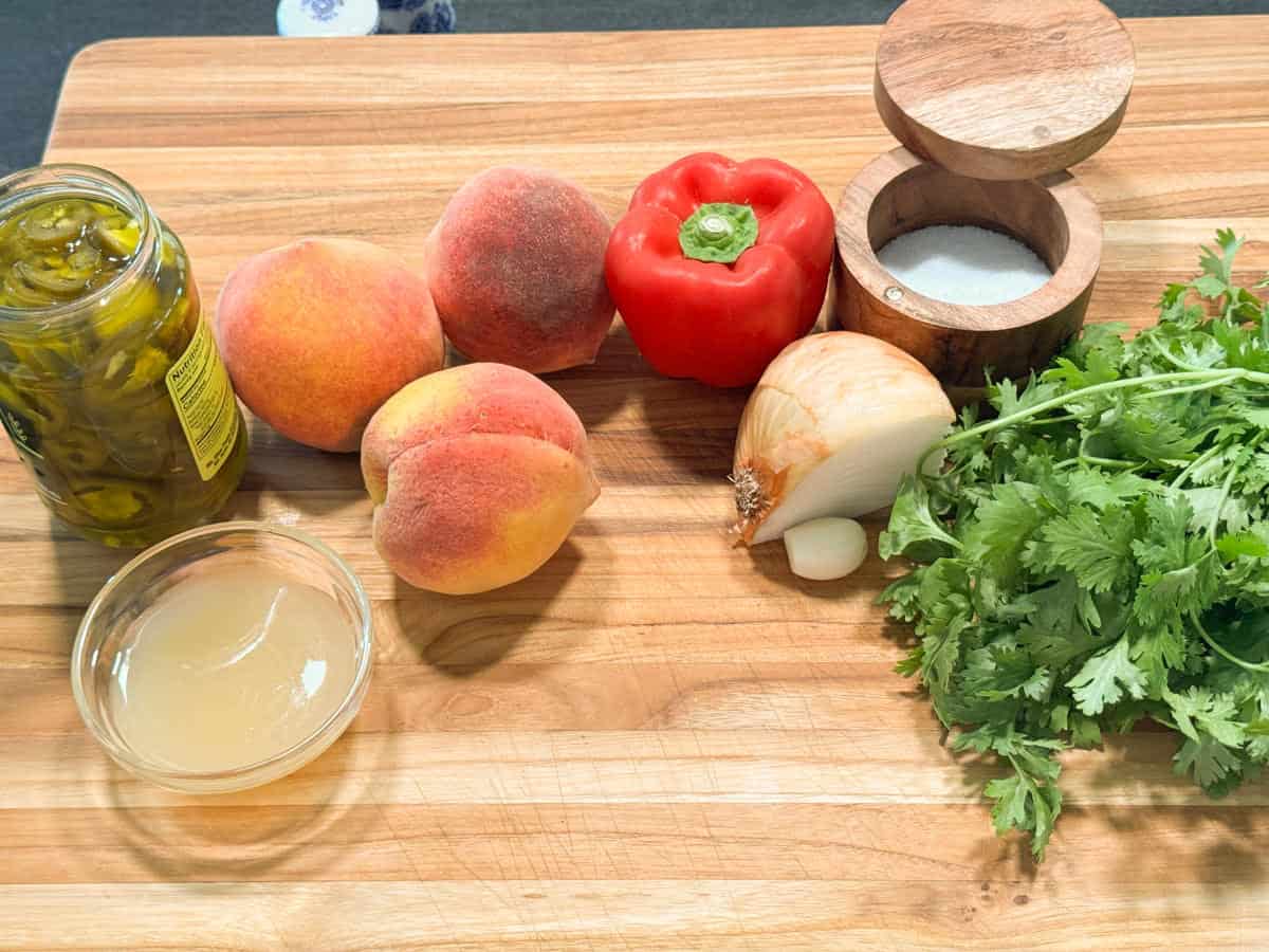 A wooden surface with a jar of pickled jalapeños, three peaches, a red bell pepper, half an onion, a bowl of liquid, a bunch of cilantro, and a wooden container of salt.