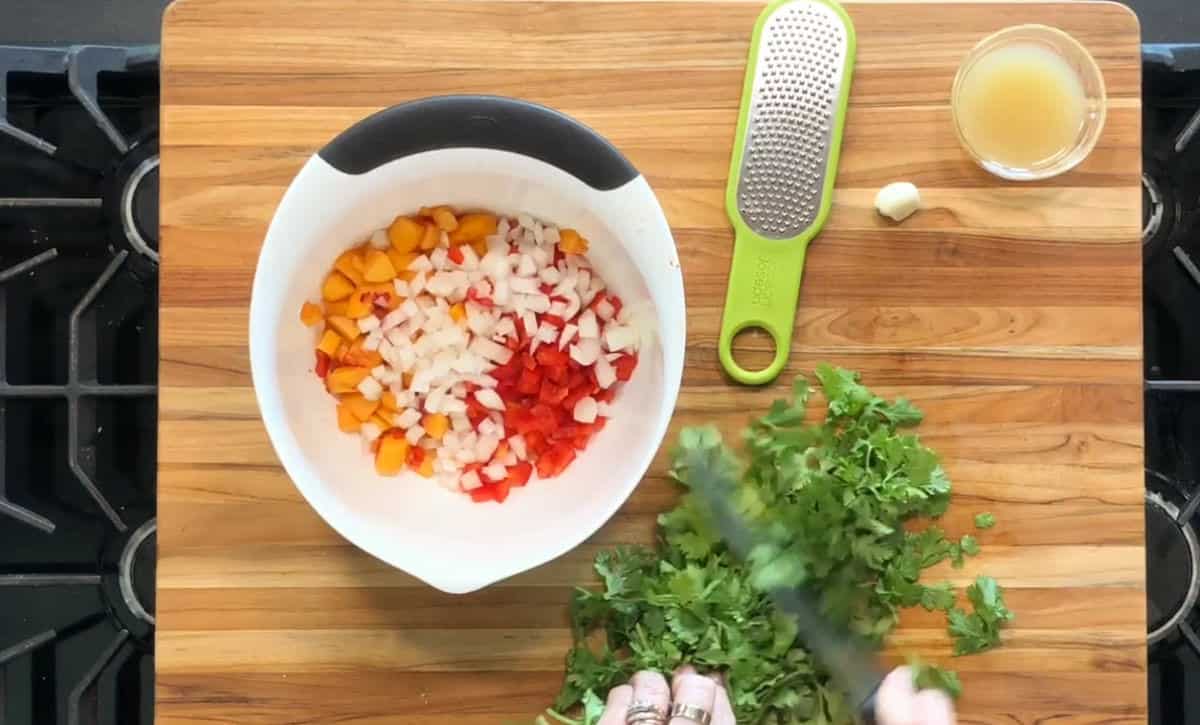 A white mixing bowl with diced yellow bell pepper, onion, and red bell pepper sits on a wooden cutting board. Nearby are chopped cilantro, a green grater, a garlic clove, and a small bowl of juice.