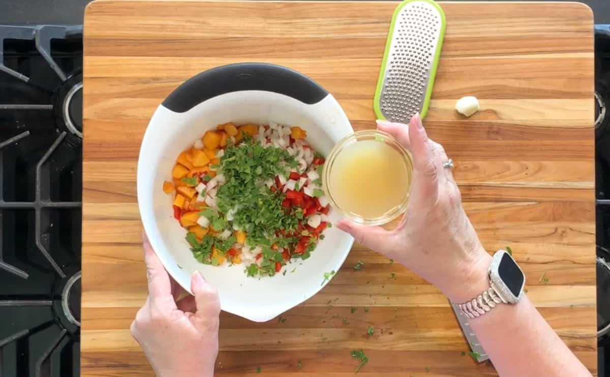 A person is holding a bowl with chopped vegetables and herbs, while pouring a small bowl of liquid into it. A grater and a garlic clove are on the wooden cutting board nearby.