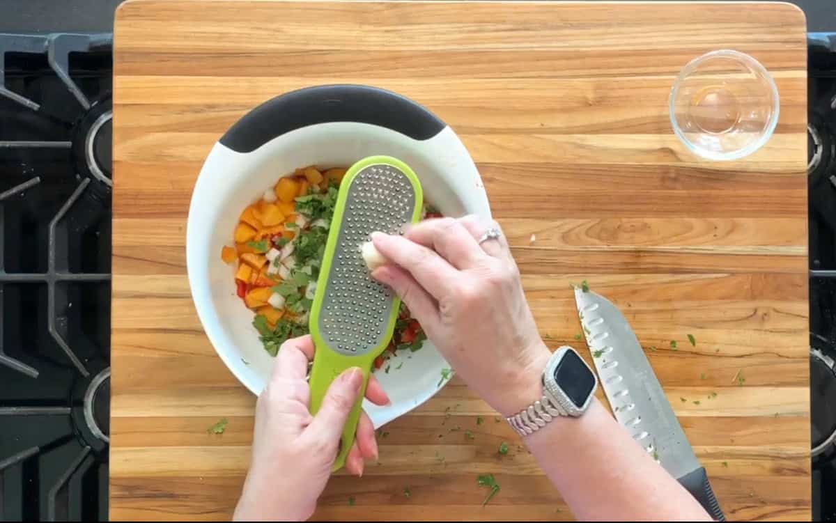 A person grates a small white ingredient, possibly garlic, into a bowl containing chopped vegetables on a wooden cutting board. A knife and an empty glass bowl are nearby. The person is wearing a watch.