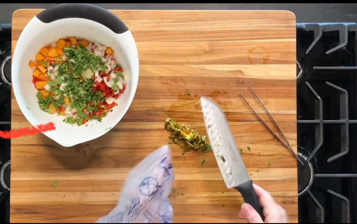A person chops herbs on a wooden cutting board next to a bowl containing chopped vegetables and herbs. Metal tongs and a gas stove are visible on the right side of the image.