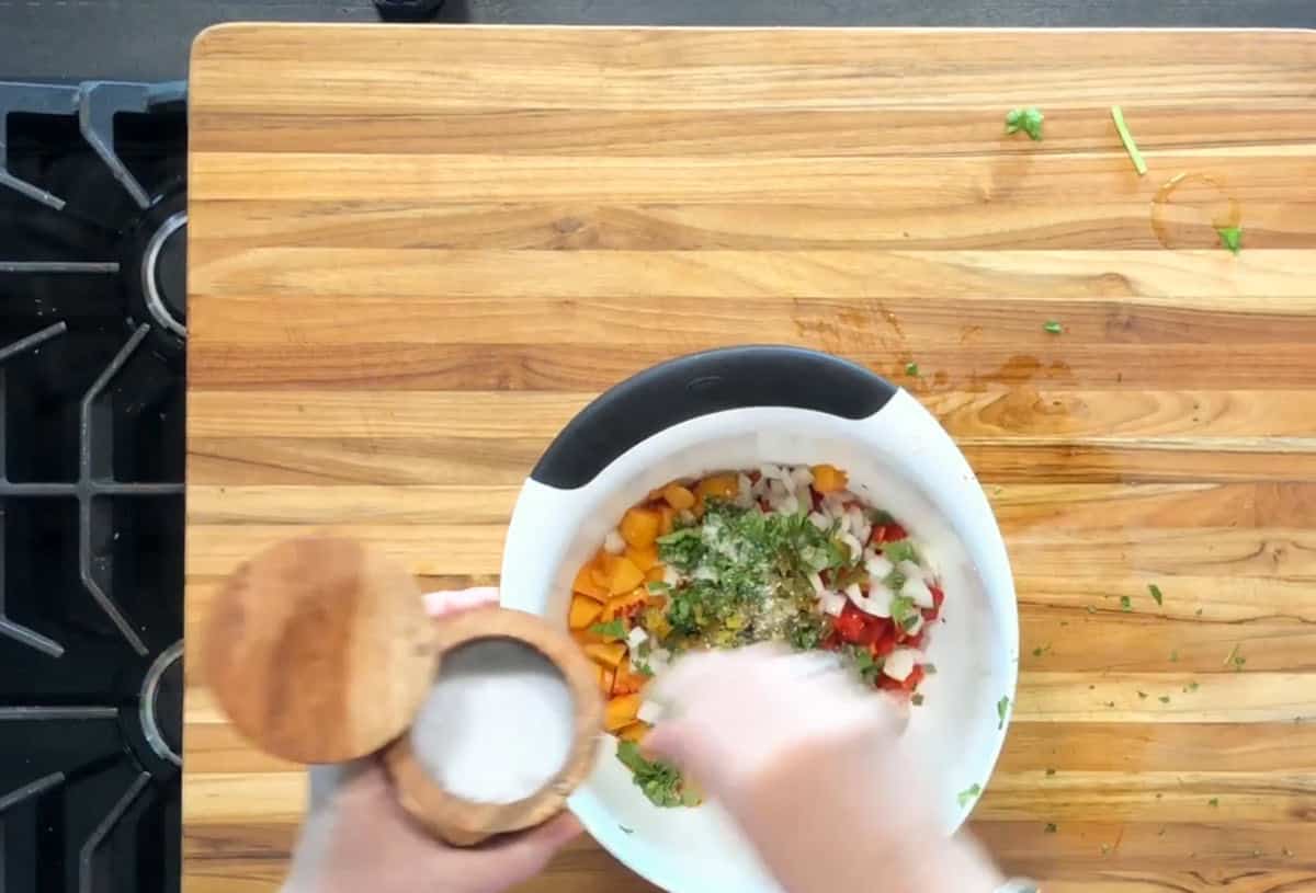 A person sprinkles salt into a white mixing bowl filled with chopped vegetables and herbs on a wooden cutting board next to a stove.