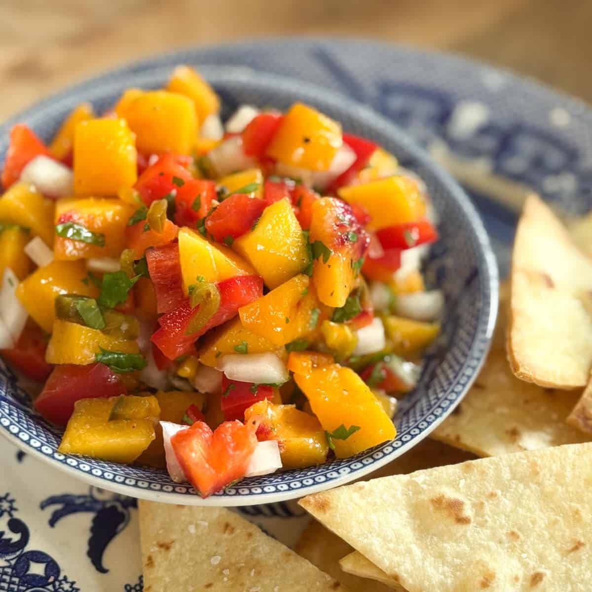 A bowl of mango salsa with diced mango, red bell pepper, onion, and herbs sits on a blue-patterned plate next to several pieces of tortilla chips.