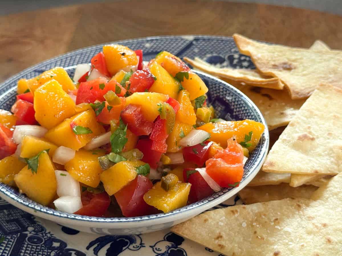A bowl of mango salsa with diced mango, tomato, onion, cilantro, and red pepper sits on a blue and white plate, next to a stack of tortilla chips on a wooden surface.